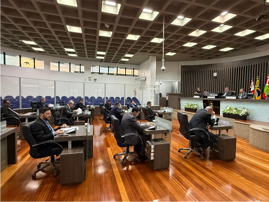 Fotografia do plenário da Câmara de Vereadores de Araranguá durante a segunda sessão ordinária de 2026. A imagem mostra os parlamentares sentados em suas bancadas individuais de madeira, organizadas em semicírculo, focados em documentos e equipamentos eletrônicos. Ao fundo, destaca-se a mesa diretora sob uma parede com painéis verticais de madeira e as bandeiras oficiais. O ambiente possui iluminação moderna embutida em um teto quadriculado e piso de madeira polida, refletindo o caráter solene da votação de projetos de saúde e infraestrutura.