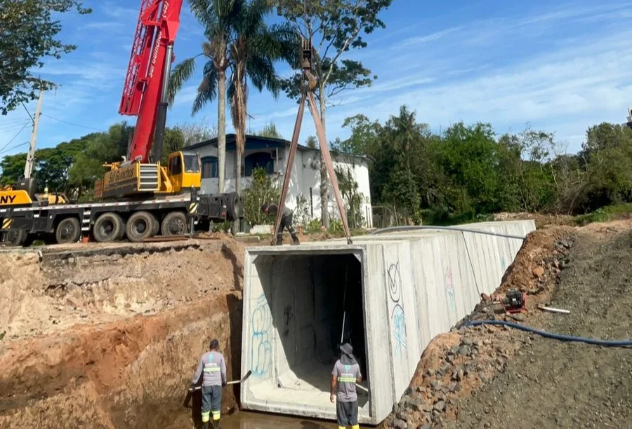 Canteiro de obras movimentado onde um grande guindaste vermelho e amarelo posiciona uma estrutura retangular de concreto pré-moldado dentro de uma vala profunda para instalação de drenagem. Trabalhadores acompanham a operação no solo. A imagem ilustra a magnitude das obras de infraestrutura que antecedem a pavimentação final da via.