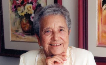 Araranguá realiza 24ª edição do Troféu Alzira Rabelo Elias Retrato de Alzira Rabelo Elias, conhecida como Dona Zizinha. A imagem mostra uma mulher idosa de cabelos grisalhos curtos e ondulados, sorrindo gentilmente para a câmera. Ela está com a mão apoiada sob o queixo, veste uma blusa clara com detalhes em renda e, ao fundo, é possível ver quadros emoldurados, incluindo uma pintura de flores.