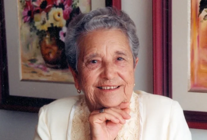 Araranguá realiza 24ª edição do Troféu Alzira Rabelo Elias Retrato de Alzira Rabelo Elias, conhecida como Dona Zizinha. A imagem mostra uma mulher idosa de cabelos grisalhos curtos e ondulados, sorrindo gentilmente para a câmera. Ela está com a mão apoiada sob o queixo, veste uma blusa clara com detalhes em renda e, ao fundo, é possível ver quadros emoldurados, incluindo uma pintura de flores.