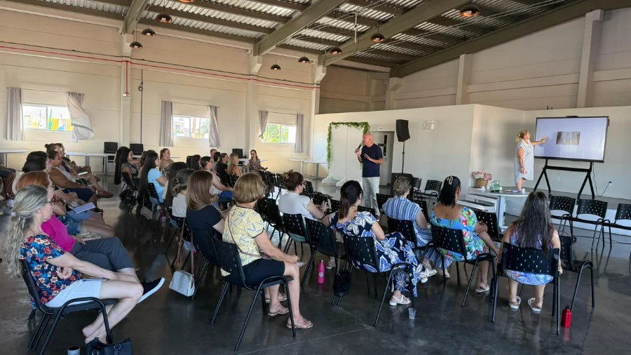 Vista ampla do salão onde ocorre o encontro de capacitação para profissionais da educação de Balneário Arroio do Silva. Uma plateia está sentada em cadeiras de frente para uma área onde um homem fala ao microfone e uma mulher aponta para uma tela de projeção.