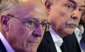 BNDES usará bilhões para enfrentar tarifas dos EUA O vice-presidente da República e ministro do Desenvolvimento, Geraldo Alckmin, aparece em primeiro plano à esquerda, com expressão séria e usando óculos. Ao fundo, à direita, o presidente do BNDES, Aloizio Mercadante, acompanha o pronunciamento durante evento realizado na capital paulista para anunciar o plano Brasil Soberano 2.0. Ambos vestem trajes formais em um ambiente de coletiva de imprensa.