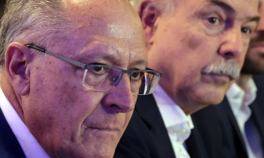 BNDES usará bilhões para enfrentar tarifas dos EUA O vice-presidente da República e ministro do Desenvolvimento, Geraldo Alckmin, aparece em primeiro plano à esquerda, com expressão séria e usando óculos. Ao fundo, à direita, o presidente do BNDES, Aloizio Mercadante, acompanha o pronunciamento durante evento realizado na capital paulista para anunciar o plano Brasil Soberano 2.0. Ambos vestem trajes formais em um ambiente de coletiva de imprensa.