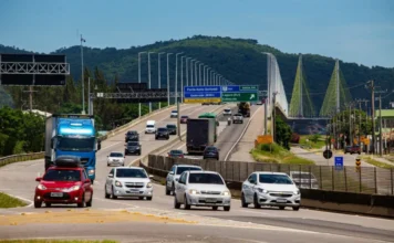 Balanço do Carnaval: BR-101 Sul recebe 1 milhão de veículos Vista em perspectiva da rodovia BR-101 Sul em Santa Catarina, mostrando o fluxo intenso de carros, caminhões e ônibus. Ao fundo, destaca-se a estrutura da Ponte Anita Garibaldi, em Laguna, sob um céu claro. A imagem ilustra a movimentação de veículos durante a Operação Carnaval da ViaCosteira.