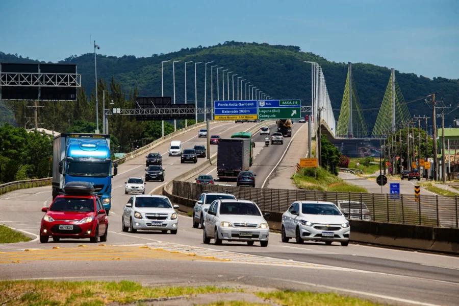 Vista em perspectiva da rodovia BR-101 Sul em Santa Catarina, mostrando o fluxo intenso de carros, caminhões e ônibus. Ao fundo, destaca-se a estrutura da Ponte Anita Garibaldi, em Laguna, sob um céu claro. A imagem ilustra a movimentação de veículos durante a Operação Carnaval da ViaCosteira.