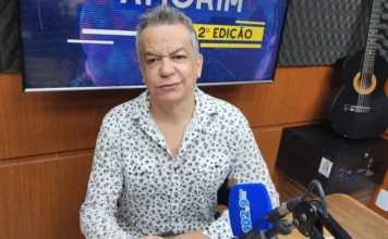 Beno César detalha trajetória e mudança para Santa Catarina Fotografia em plano médio do cantor e produtor musical Beno César, um homem de pele clara e cabelos grisalhos penteados para trás, vestindo uma camisa branca com estampa floral preta. Ele está sentado em uma mesa de estúdio de rádio, posicionado atrás de um microfone azul com a marca 102.9 Amorim FM. Ao fundo, um monitor exibe a identidade visual do Jornal Amorim 2ª Edição e, à direita, um violão preto está pendurado na parede com isolamento acústico.