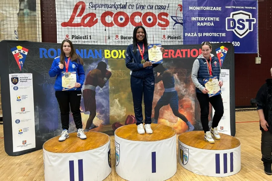 A imagem mostra a boxeadora Carolina de Vargas Pires da Silva no centro de um pódio circular de madeira, ocupando o primeiro lugar. Ela é uma jovem negra, está sorrindo, veste um agasalho esportivo azul-marinho da Seleção Brasileira e segura um certificado de premiação, exibindo a medalha de ouro no peito. À sua esquerda e direita, nos degraus de segundo e terceiro lugar, estão outras duas competidoras segurando seus certificados. Ao fundo, há um grande banner da Federação Romena de Boxe (Romanian Boxing Federation) com ilustrações de atletas em combate e logotipos de patrocinadores locais.