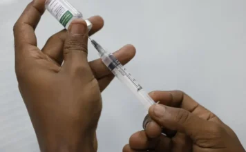 Brasil registra 29 mortes por Covid-19 no início do ano Mãos de uma pessoa preparando uma vacina, com uma seringa e um frasco, ilustrando o processo de imunização contra a Covid-19 e outras doenças respiratórias, tema central da matéria sobre a importância da vacinação e a baixa adesão no Brasil.