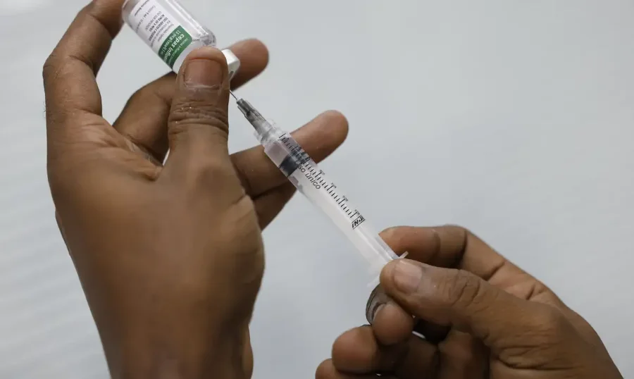 Mãos de uma pessoa preparando uma vacina, com uma seringa e um frasco, ilustrando o processo de imunização contra a Covid-19 e outras doenças respiratórias, tema central da matéria sobre a importância da vacinação e a baixa adesão no Brasil.