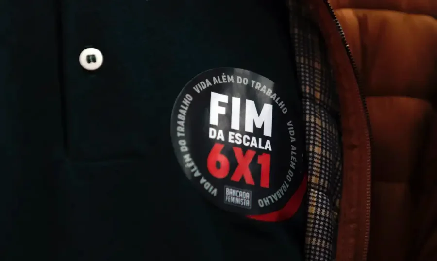 Close de um adesivo circular fixado em uma peça de roupa escura. O adesivo possui fundo preto com bordas brancas, onde se lê a frase repetida 