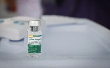 Butantan antecipa 1,3 milhão de vacinas contra dengue Fotografia em close-up de um frasco de vidro da vacina Butantan-DV contra a dengue. O rótulo, com detalhes em verde e branco, identifica o imunizante e o logotipo do Instituto Butantan. O frasco está posicionado sobre uma superfície branca em um ambiente clínico, com o fundo levemente desfocado.