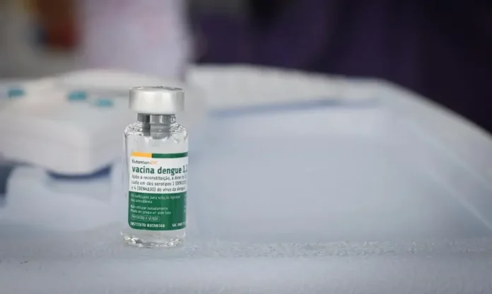 Butantan antecipa 1,3 milhão de vacinas contra dengue Fotografia em close-up de um frasco de vidro da vacina Butantan-DV contra a dengue. O rótulo, com detalhes em verde e branco, identifica o imunizante e o logotipo do Instituto Butantan. O frasco está posicionado sobre uma superfície branca em um ambiente clínico, com o fundo levemente desfocado.