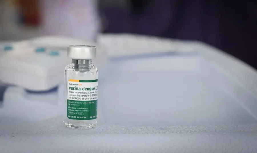 Butantan antecipa 1,3 milhão de vacinas contra dengue Fotografia em close-up de um frasco de vidro da vacina Butantan-DV contra a dengue. O rótulo, com detalhes em verde e branco, identifica o imunizante e o logotipo do Instituto Butantan. O frasco está posicionado sobre uma superfície branca em um ambiente clínico, com o fundo levemente desfocado.