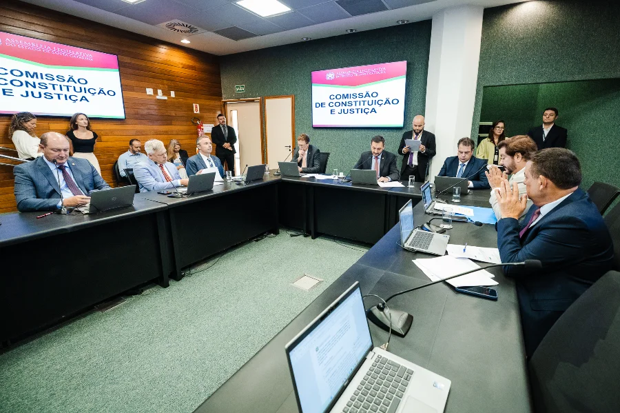 Foto da primeira reunião da Comissão de Constituição e Justiça (CCJ) da Assembleia Legislativa de Santa Catarina (ALESC) em 2026, realizada em um auditório. Vários deputados e assessores estão sentados ao redor de uma grande mesa de conferência, com laptops e documentos. No centro da mesa, um deputado preside a sessão. Telas na parede de fundo exibem o logotipo da ALESC e o nome da comissão. A reunião abordou a aprovação de projetos como o Cordão de Girassol para deficiências ocultas.