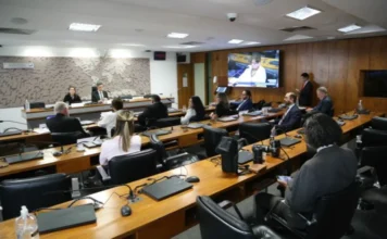 CCS elege nova presidência e empossa membros nesta segunda Fotografia colorida do interior do Plenário 3 da ala Alexandre Costa, no Senado Federal. A imagem mostra diversos conselheiros do CCS sentados em bancadas de madeira dispostas em fileiras, equipadas com microfones e laptops. Ao fundo, há uma parede com um mapa-múndi estilizado e um monitor de TV exibindo a transmissão da sessão. O ambiente é formal e bem iluminado.
