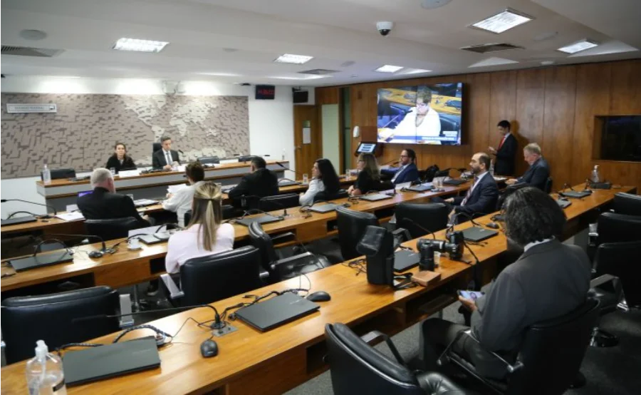 Fotografia colorida do interior do Plenário 3 da ala Alexandre Costa, no Senado Federal. A imagem mostra diversos conselheiros do CCS sentados em bancadas de madeira dispostas em fileiras, equipadas com microfones e laptops. Ao fundo, há uma parede com um mapa-múndi estilizado e um monitor de TV exibindo a transmissão da sessão. O ambiente é formal e bem iluminado.