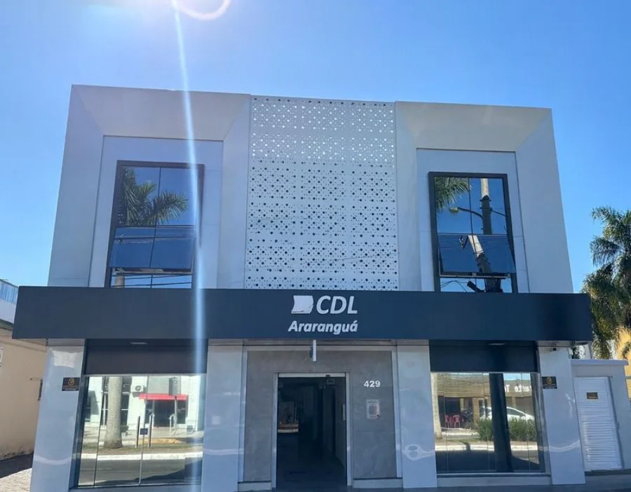 Fachada frontal da sede da Câmara de Dirigentes Lojistas (CDL) de Araranguá, em Santa Catarina. O prédio de arquitetura moderna apresenta janelas espelhadas e uma placa preta centralizada com o logotipo da entidade em letras brancas. A imagem ilustra a instituição que lidera o movimento de preocupação com os impactos econômicos do novo projeto de lei estadual.