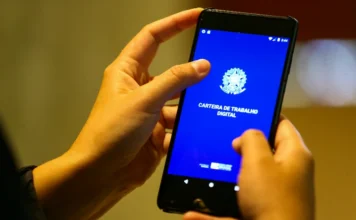 COFEM alerta para impactos da jornada de 36 horas em SC Pessoa segurando um smartphone que exibe a tela inicial do aplicativo da Carteira de Trabalho Digital. A imagem ilustra o debate sobre as mudanças na jornada de trabalho e seus possíveis efeitos na formalidade do emprego no Brasil.