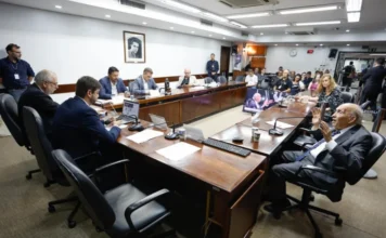 CPI das Rodovias debate custos e sistema Free Flow no RS Reunião da CPI dos Contratos de Concessão das Rodovias Estaduais na Assembleia Legislativa do Rio Grande do Sul. O conselheiro do DAER, Luís Roberto Ponte, um homem idoso de terno e gravata, gesticula com as mãos enquanto presta depoimento aos deputados sentados ao redor de uma longa mesa de madeira em uma sala de comissões. Vários parlamentares e assessores observam, e uma tela ao fundo exibe uma imagem da reunião.