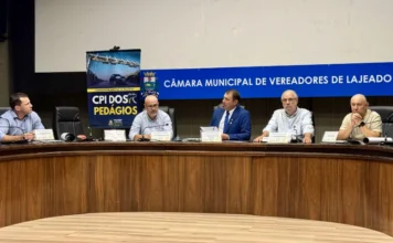 CPI debate custos do novo pedágio em audiência em Lajeado Fotografia horizontal mostra cinco homens sentados à mesa diretora durante a audiência pública da CPI dos Pedágios na Câmara Municipal de Lajeado. Eles estão posicionados atrás de uma bancada semicircular de madeira escura. Ao fundo, na parede cinza, destaca-se um painel azul com a inscrição "Câmara Municipal de Vereadores de Lajeado" em letras brancas. À esquerda, há um banner vertical da "CPI dos Pedágios" com a imagem de um pórtico de sistema free flow. Os participantes vestem camisas sociais e paletós, e sobre a mesa há microfones, garrafas de água e placas de identificação.