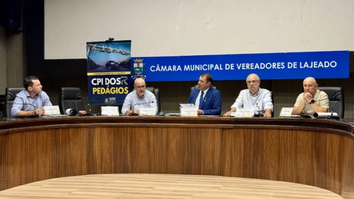 CPI debate custos do novo pedágio em audiência em Lajeado Fotografia horizontal mostra cinco homens sentados à mesa diretora durante a audiência pública da CPI dos Pedágios na Câmara Municipal de Lajeado. Eles estão posicionados atrás de uma bancada semicircular de madeira escura. Ao fundo, na parede cinza, destaca-se um painel azul com a inscrição "Câmara Municipal de Vereadores de Lajeado" em letras brancas. À esquerda, há um banner vertical da "CPI dos Pedágios" com a imagem de um pórtico de sistema free flow. Os participantes vestem camisas sociais e paletós, e sobre a mesa há microfones, garrafas de água e placas de identificação.