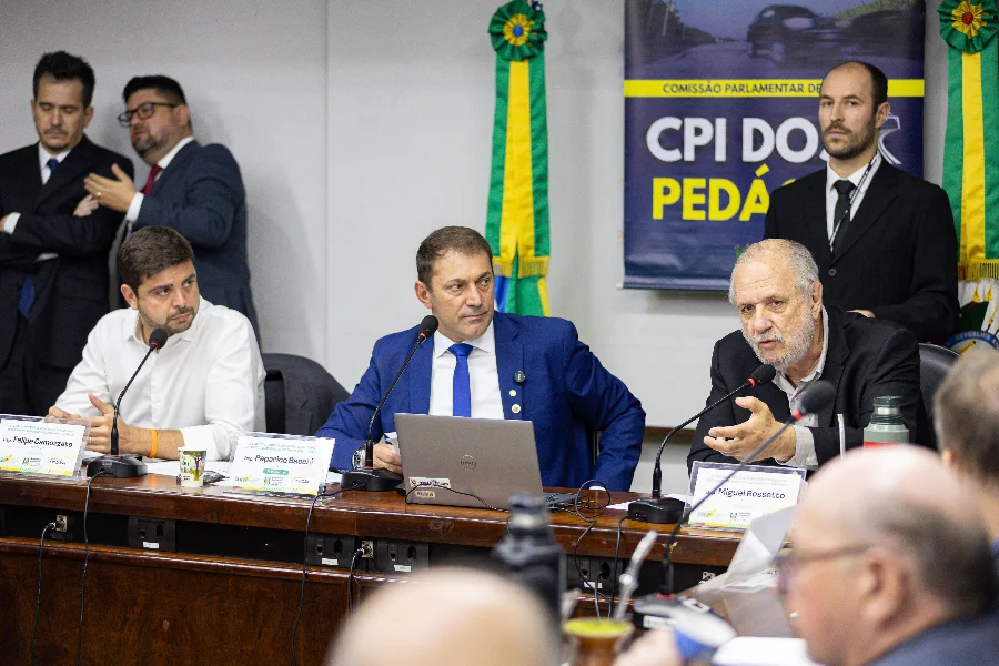 Deputados participam de reunião da CPI dos Pedágios na Assembleia Legislativa do Rio Grande do Sul. O deputado Miguel Rossetto (à direita), relator da comissão, fala ao microfone e gesticula, observado pelos deputados Paparico Bacchi (ao centro) e Felipe Camozzato (à esquerda). Ao fundo, vê-se um banner com a inscrição 