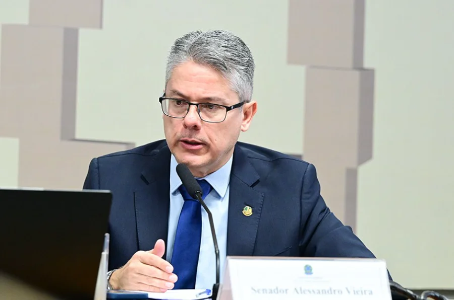 Fotografia em plano médio do senador Alessandro Vieira, relator da CPI do Crime Organizado. Ele é um homem de pele clara, cabelos grisalhos e usa óculos de grau. Está vestido com um terno azul marinho e gravata azul, falando ao microfone durante uma sessão oficial no Senado Federal. À sua frente, há uma placa de identificação com seu nome.