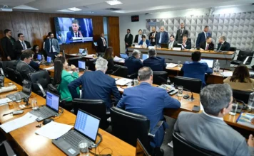 CPMI do INSS: Ingrid Santos substitui Daniel Vorcaro Fotografia em ângulo aberto de uma sala de comissões no Senado Federal durante a reunião da CPMI do INSS. Parlamentares e técnicos estão sentados em mesas de madeira dispostas em semicírculo, com diversos laptops abertos. Ao fundo, uma tela de televisão exibe a transmissão ao vivo de um orador. À direita, integrantes da mesa diretora coordenam os trabalhos em uma bancada elevada, enquanto o ambiente é ocupado por assessores e jornalistas.