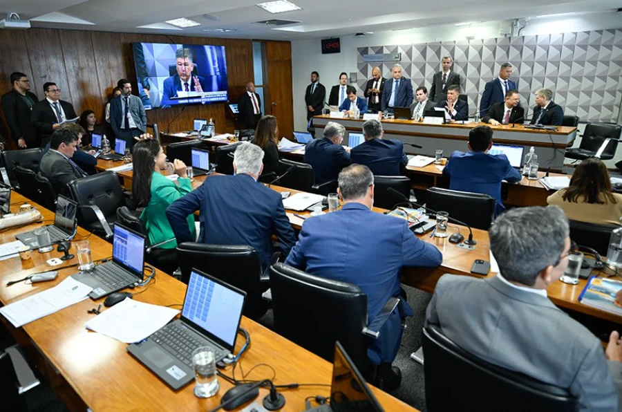 CPMI do INSS Ingrid Santos substitui Daniel Vorcaro Fotografia em ângulo aberto de uma sala de comissões no Senado Federal durante a reunião da CPMI do INSS. Parlamentares e técnicos estão sentados em mesas de madeira dispostas em semicírculo, com diversos laptops abertos. Ao fundo, uma tela de televisão exibe a transmissão ao vivo de um orador. À direita, integrantes da mesa diretora coordenam os trabalhos em uma bancada elevada, enquanto o ambiente é ocupado por assessores e jornalistas.