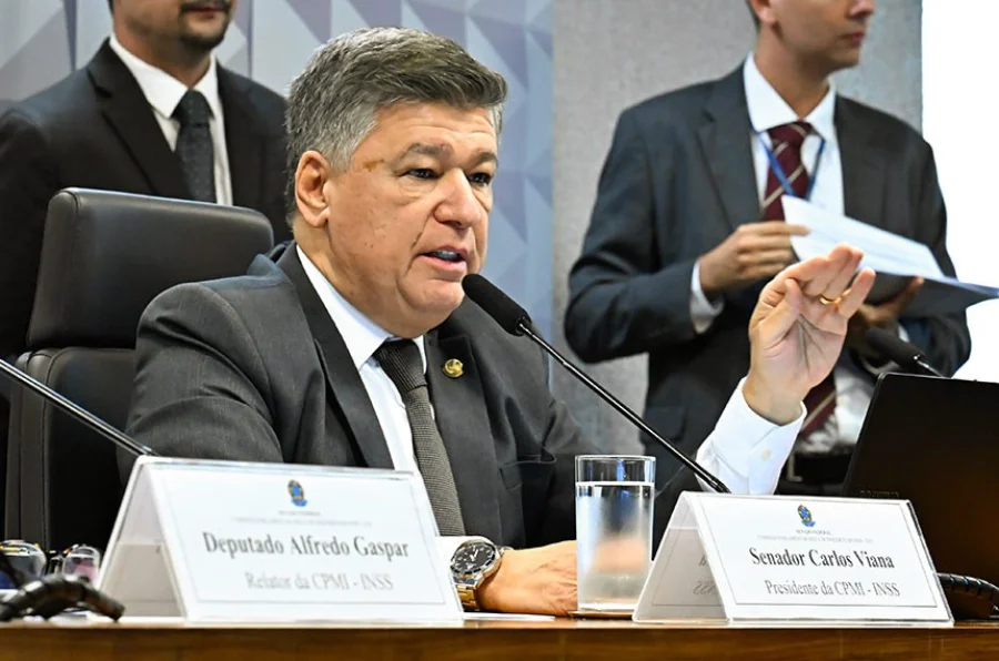 O senador Carlos Viana, presidente da CPMI do INSS, está sentado à mesa diretora da comissão falando ao microfone. Ele veste terno cinza e gesticula com a mão esquerda enquanto fala. À sua frente, veem-se placas de identificação com os nomes do relator, deputado Alfredo Gaspar, e o dele próprio.
