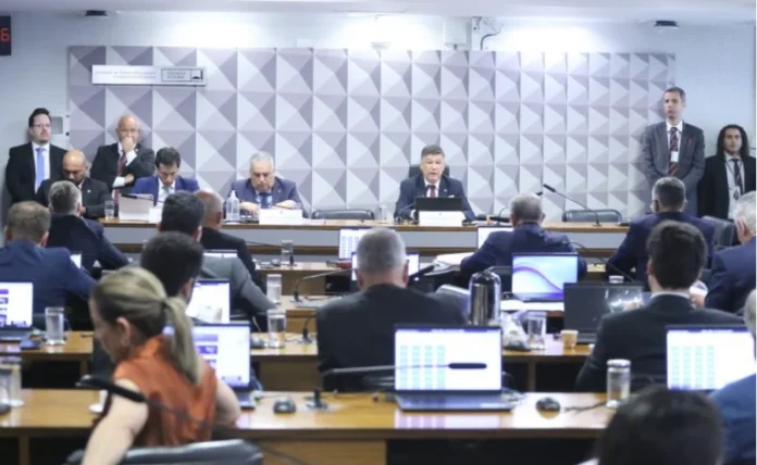 CPMI do INSS convoca Ingrid Pikinskeni para depor no Senado Fotografia em plano médio de uma reunião parlamentar no Senado Federal. Ao fundo, cinco parlamentares homens vestindo ternos escuros estão sentados atrás de uma bancada de madeira elevada. À frente deles, em primeiro plano, diversas pessoas aparecem de costas, sentadas em bancadas com notebooks abertos, acompanhando a sessão. A parede ao fundo possui um painel decorativo com padrões geométricos em tons de cinza. O ambiente é formal e bem iluminado.