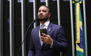 Câmara analisa novas regras para mandados de busca em casas Fotografia em plano médio do deputado federal Capitão Alden, autor do Projeto de Lei 6480/25, discursando na tribuna da Câmara dos Deputados. O parlamentar, um homem pardo de barba e cabelos curtos, veste terno azul-marinho, camisa branca e gravata clara. Ele segura um smartphone na mão esquerda enquanto fala ao microfone. Ao fundo, é possível observar as listras verticais do plenário e parte da bandeira do Brasil.