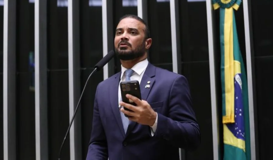 Câmara analisa novas regras para mandados de busca em casas Fotografia em plano médio do deputado federal Capitão Alden, autor do Projeto de Lei 6480/25, discursando na tribuna da Câmara dos Deputados. O parlamentar, um homem pardo de barba e cabelos curtos, veste terno azul-marinho, camisa branca e gravata clara. Ele segura um smartphone na mão esquerda enquanto fala ao microfone. Ao fundo, é possível observar as listras verticais do plenário e parte da bandeira do Brasil.