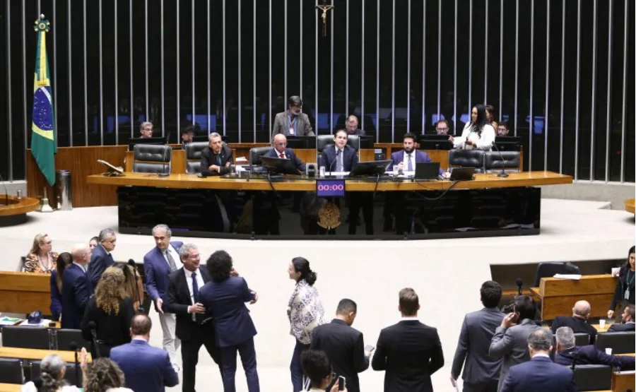 A imagem mostra uma vista geral do Plenário da Câmara dos Deputados durante uma sessão de votação. No centro da composição, a mesa diretora é ocupada por parlamentares, com o painel eletrônico de votação exibindo 