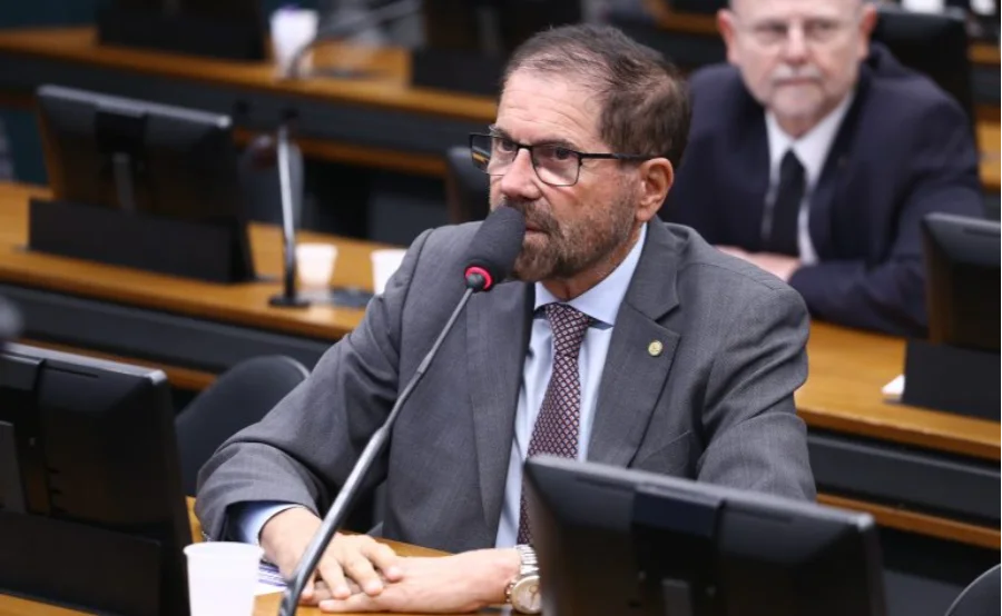 O deputado federal José Rocha (União-BA), relator do projeto, aparece sentado à mesa de uma comissão na Câmara dos Deputados. Ele é um homem de pele clara, com barba e cabelos castanhos curtos, usa óculos de armação escura, terno cinza e gravata estampada. Ele fala ao microfone em um ambiente legislativo, com monitores e outras autoridades ao fundo em uma composição desfocada.