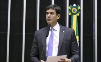 Câmara avalia leilão de bens em pátios policiais Fotografia em plano médio do deputado Rafael Prudente (MDB-DF) discursando na tribuna da Câmara dos Deputados. Ele veste terno escuro, camisa branca e gravata lilás, segurando papéis enquanto fala ao microfone. Ao fundo, as colunas verticais do plenário e uma bandeira do Brasil. A imagem ilustra o autor da proposta que visa regularizar o destino de bens e veículos abandonados em pátios policiais.