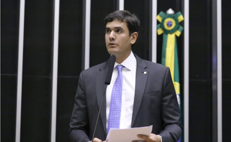 Câmara avalia leilão de bens em pátios policiais Fotografia em plano médio do deputado Rafael Prudente (MDB-DF) discursando na tribuna da Câmara dos Deputados. Ele veste terno escuro, camisa branca e gravata lilás, segurando papéis enquanto fala ao microfone. Ao fundo, as colunas verticais do plenário e uma bandeira do Brasil. A imagem ilustra o autor da proposta que visa regularizar o destino de bens e veículos abandonados em pátios policiais.