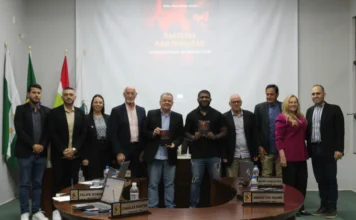 Câmara de Forquilhinha debate igualdade racial em sessão Fotografia em grupo no plenário da Câmara de Vereadores de Forquilhinha. Ao centro, o autor Sérgio Nunes Vitório Júnior e um parlamentar seguram exemplares do livro "Racistas não passarão", que possui capa escura com detalhes em vermelho. Ao fundo, uma tela de projeção exibe o título da obra e o subtítulo "O ódio colonial da branquitude". O grupo, composto por dez pessoas entre homens e mulheres em trajes formais, posa atrás da bancada principal, onde se veem as placas de identificação dos vereadores. À esquerda, as bandeiras do Brasil, de Santa Catarina e do município compõem o cenário legislativo.