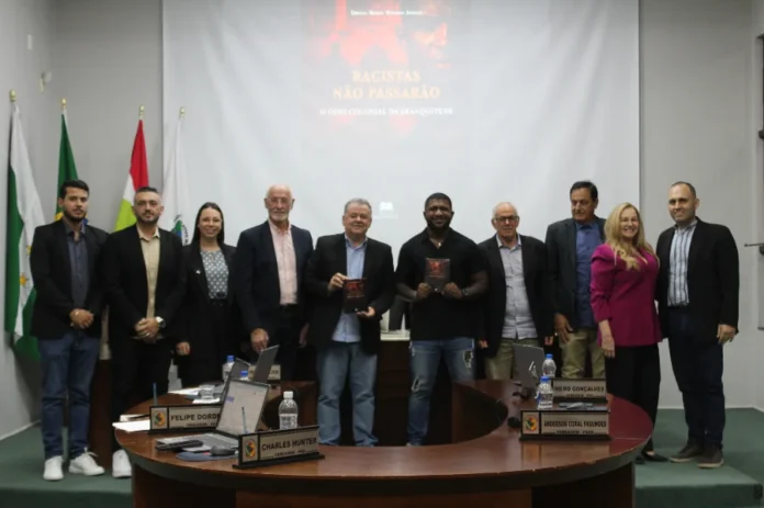 Câmara de Forquilhinha debate igualdade racial em sessão Fotografia em grupo no plenário da Câmara de Vereadores de Forquilhinha. Ao centro, o autor Sérgio Nunes Vitório Júnior e um parlamentar seguram exemplares do livro "Racistas não passarão", que possui capa escura com detalhes em vermelho. Ao fundo, uma tela de projeção exibe o título da obra e o subtítulo "O ódio colonial da branquitude". O grupo, composto por dez pessoas entre homens e mulheres em trajes formais, posa atrás da bancada principal, onde se veem as placas de identificação dos vereadores. À esquerda, as bandeiras do Brasil, de Santa Catarina e do município compõem o cenário legislativo.