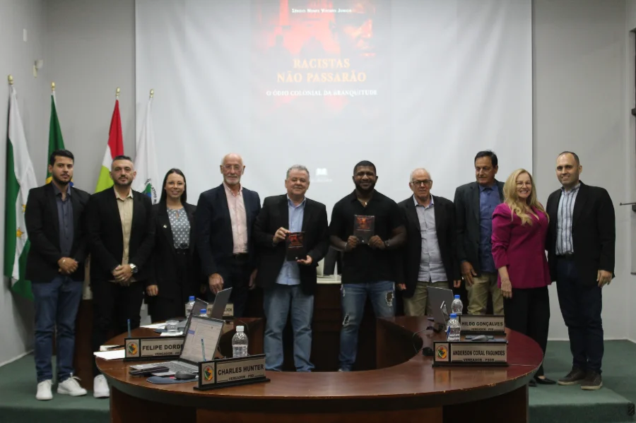 Fotografia em grupo no plenário da Câmara de Vereadores de Forquilhinha. Ao centro, o autor Sérgio Nunes Vitório Júnior e um parlamentar seguram exemplares do livro 