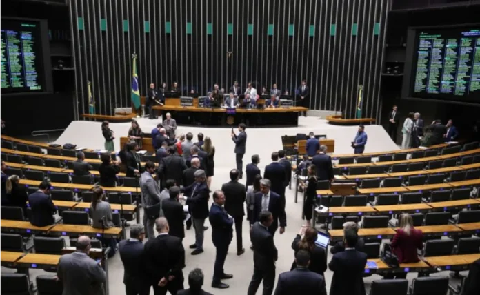 Câmara inicia votações com Gás do Povo e crédito rural Vista geral do interior do Plenário da Câmara dos Deputados, com vários parlamentares de terno em pé conversando no centro e a mesa diretora ocupada ao fundo. Telões eletrônicos nas paredes laterais exibem informações em verde. A imagem ilustra o local onde ocorrerá a primeira sessão de votações do ano para analisar as medidas provisórias do Programa Gás do Povo e do crédito rural.