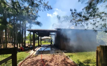 Campanha arrecada fraldas e roupas após incêndio em Sombrio Registro do local do incêndio que destruiu uma residência de madeira no bairro Januária, em Sombrio. A imagem mostra a atuação de um bombeiro e a fumaça saindo da estrutura, evidenciando o incidente que resultou na perda total dos bens da família, que agora solicita doações de fraldas e roupas.