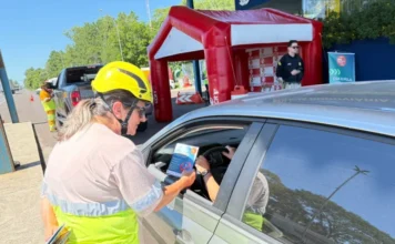 Carnaval: ViaCosteira alerta para álcool e direção Funcionária da concessionária ViaCosteira, utilizando colete reflexivo e capacete de segurança, entrega panfleto educativo a um motorista durante ação de conscientização na rodovia. Ao fundo, uma estrutura inflável vermelha e um agente de segurança acompanham a atividade.