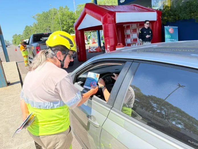 Carnaval ViaCosteira alerta para álcool e direção Funcionária da concessionária ViaCosteira, utilizando colete reflexivo e capacete de segurança, entrega panfleto educativo a um motorista durante ação de conscientização na rodovia. Ao fundo, uma estrutura inflável vermelha e um agente de segurança acompanham a atividade.