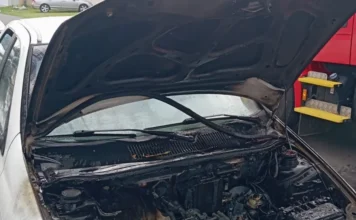 Carro pega fogo e mobiliza bombeiros em Sombrio Fotografia em plano médio de um automóvel branco com o capô levantado, revelando o compartimento do motor severamente danificado e carbonizado por um incêndio. O interior está coberto por fuligem preta e resíduos de queima. No canto direito da imagem, é visível parte de uma viatura vermelha do Corpo de Bombeiros, indicando o local da ocorrência em via pública.