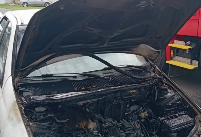 Carro pega fogo e mobiliza bombeiros em Sombrio Fotografia em plano médio de um automóvel branco com o capô levantado, revelando o compartimento do motor severamente danificado e carbonizado por um incêndio. O interior está coberto por fuligem preta e resíduos de queima. No canto direito da imagem, é visível parte de uma viatura vermelha do Corpo de Bombeiros, indicando o local da ocorrência em via pública.