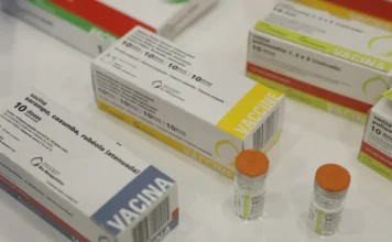 Casos de sarampo nas Américas crescem 32 vezes em um ano Fotografia em close e levemente inclinada mostrando diversas caixas e frascos de imunizantes sobre uma superfície clara. Em destaque, no primeiro plano à esquerda, aparece uma caixa branca da vacina contra sarampo, caxumba e rubéola (tríplice viral), com o selo da Bio-Manguinhos/Fiocruz. Ao fundo, outras caixas de vacinas e dois pequenos frascos de vidro com tampas laranjas completam a composição, simbolizando a estrutura de imunização necessária para o controle de surtos.