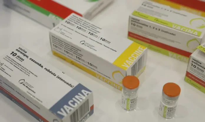 Casos de sarampo nas Américas crescem 32 vezes em um ano Fotografia em close e levemente inclinada mostrando diversas caixas e frascos de imunizantes sobre uma superfície clara. Em destaque, no primeiro plano à esquerda, aparece uma caixa branca da vacina contra sarampo, caxumba e rubéola (tríplice viral), com o selo da Bio-Manguinhos/Fiocruz. Ao fundo, outras caixas de vacinas e dois pequenos frascos de vidro com tampas laranjas completam a composição, simbolizando a estrutura de imunização necessária para o controle de surtos.