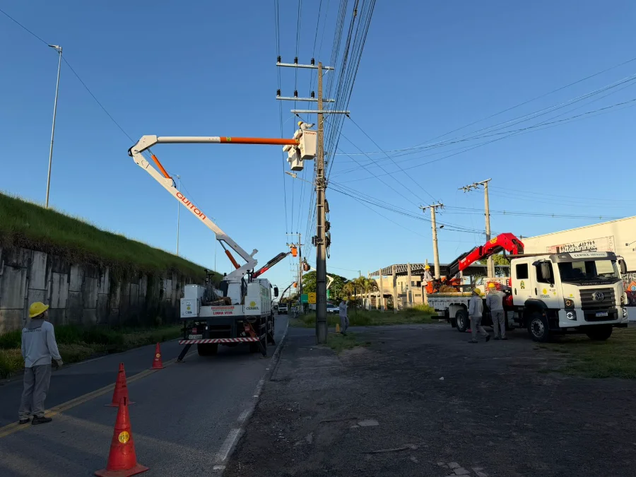 Equipes técnicas e caminhões da Cersul, incluindo um com cesto aéreo, realizam trabalhos de reforço na rede elétrica em postes de energia na beira de uma estrada, obra necessária para atender a nova demanda da escola em Araranguá.