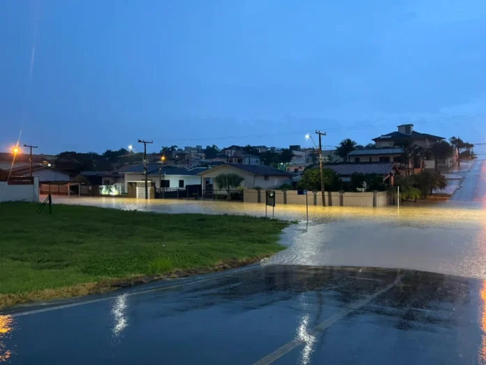 Chuva intensa alaga ruas e atinge casas em Cocal do Sul Fotografia de uma rua residencial em Cocal do Sul sob condições de alagamento ao anoitecer. A água barrenta cobre totalmente o asfalto, atingindo o nível das calçadas e muros das casas. As luzes dos postes de iluminação pública refletem na superfície da água. Ao fundo, veem-se casas em terrenos mais elevados e o céu em tons de azul escuro e cinza, indicando tempo nublado e chuvoso.